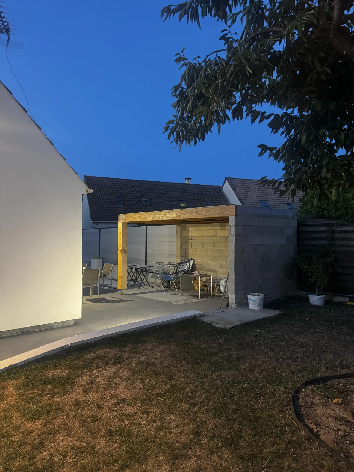 Photographie en fin de journée de l'abri couvert dans le jardin, avec sa poutre et son poteau en bois et ses murs en parpaings, à côté de la maison.